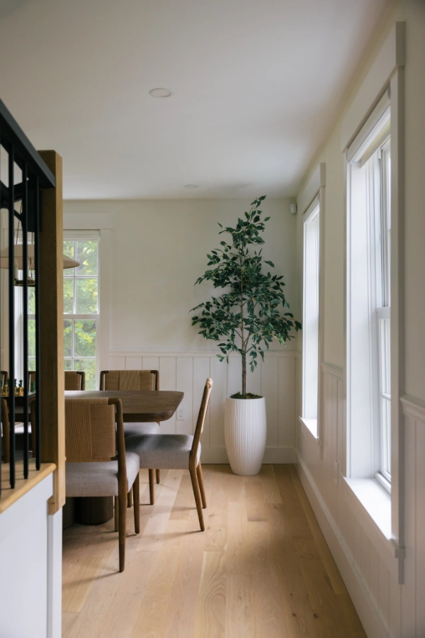 Dining area beside library shelving with upholstered chairs, light wood floors, tall windows, and minimalist plant decor